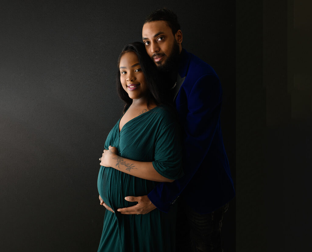 pregnant woman with her husband standing behind and cuddlling the pregnant belly. both parent-to-be are smiling and posing for the photoshoot
